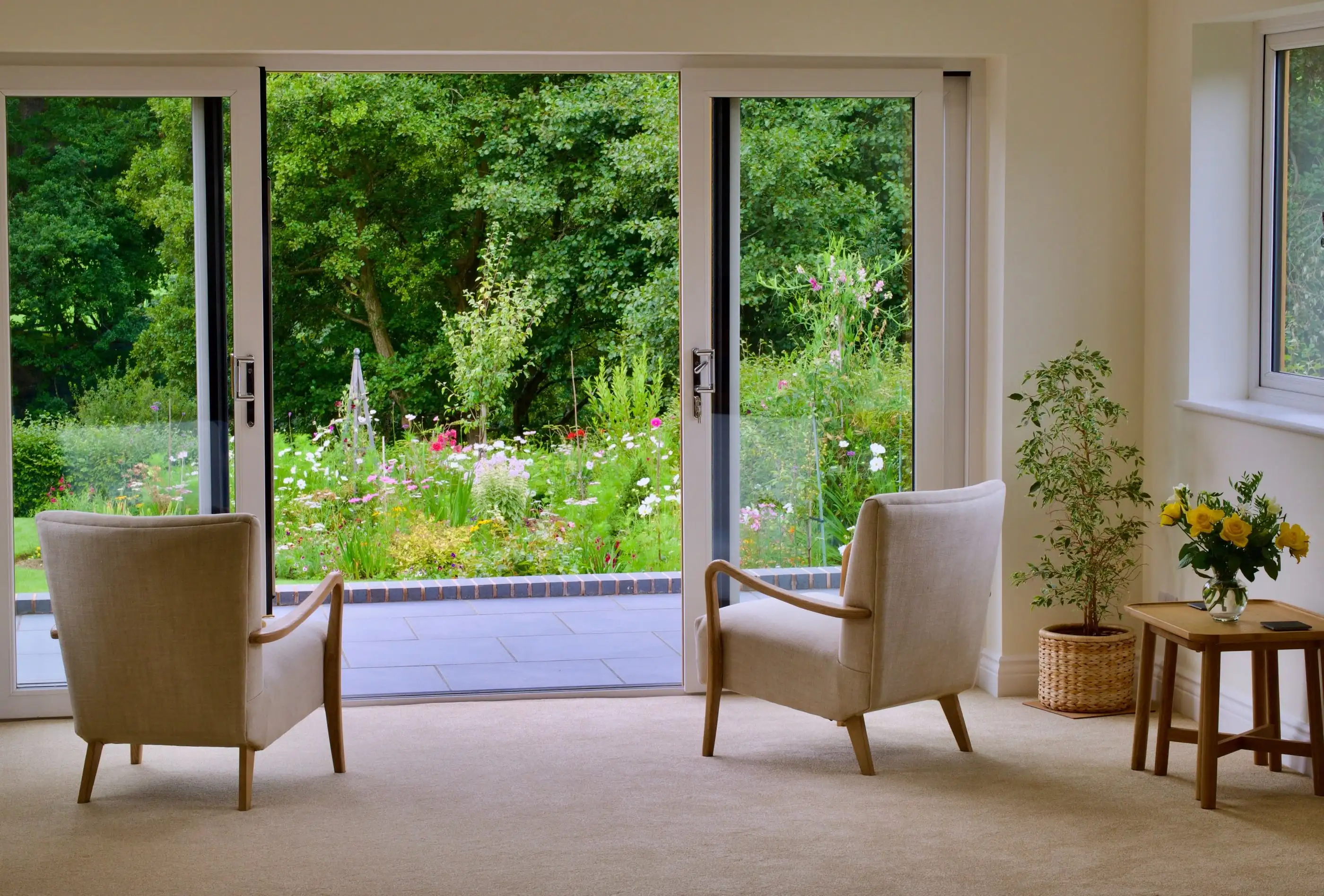 Salon aux fauteuils clairs, baie vitrée grande ouverte sur un jardin de vivaces et de grands arbres.